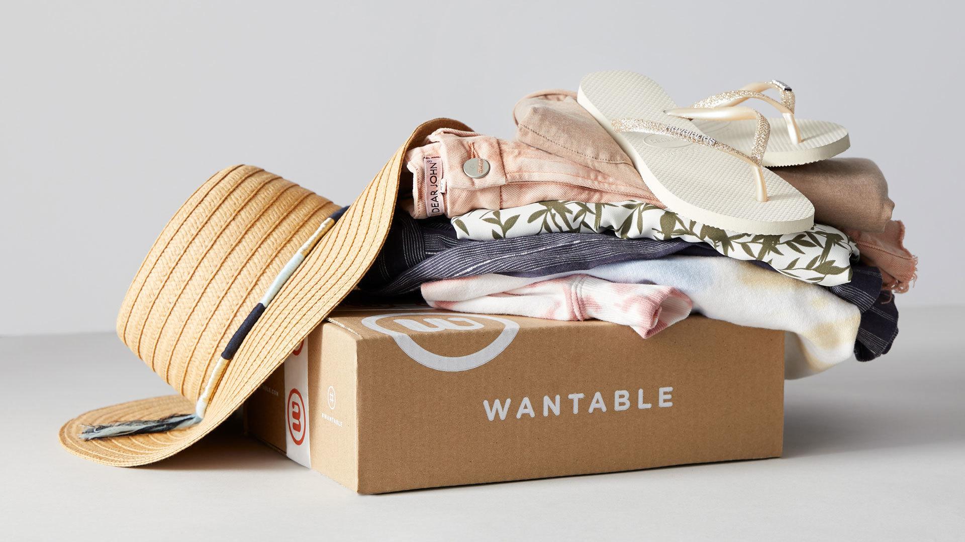 Wantable box with a stack of clothing on top topped with flip flops, off the left side is a straw bat with blue, gold, and white band