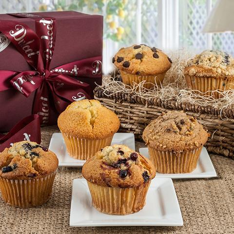 Muffins and maroon muffin box sitting on a table with a window in the background showing a lemon tree.  three of the muffins are sitting on individual square white plates, a fourth is sitting on the table, on the left of the photo is a maroon muffin box with bow and in the back is a low sided basket with decorative fill and two muffins sitting on top