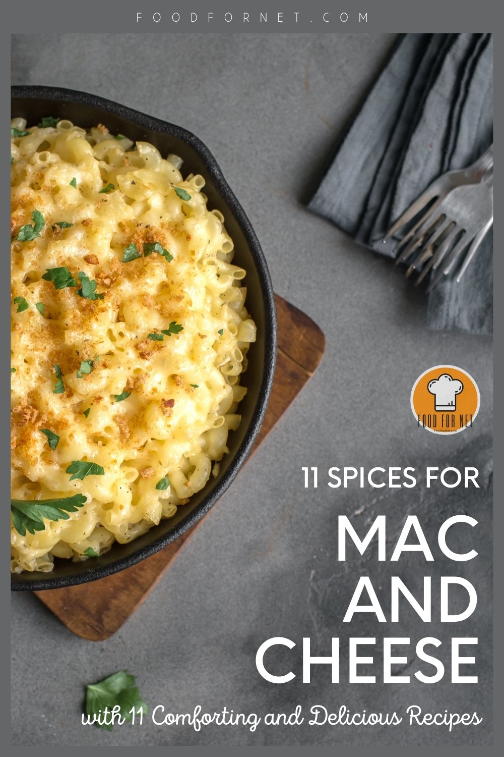 Spices for Mac and Cheese. top view image of a grey surface with a skillet pan with mac and cheese with herbs on top, resting on a wooden board, beside it is a grey-blue napkin with two silver forks 