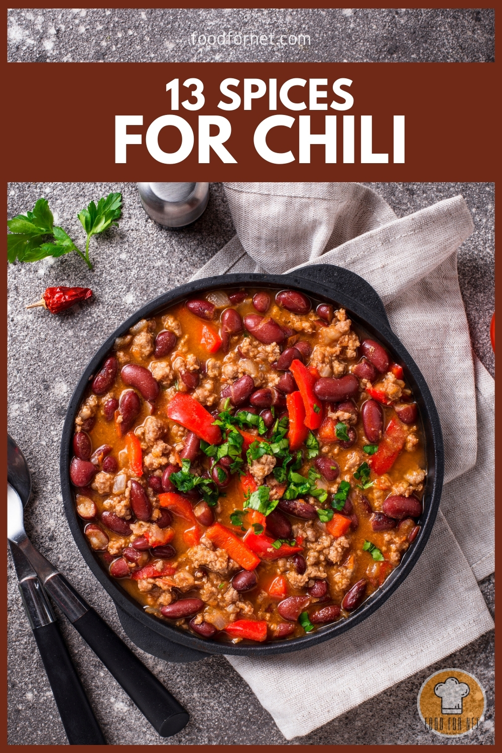 top view image of a skillet of chili con carne resting on top of a table napkin on a marble surface, with spoon and fork, salt shaker, dried chili, and fresh coriander beside it.