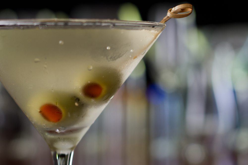 A glass containing a martini that has been garnished with two olives. An out of focus bar can be seen in the background. 