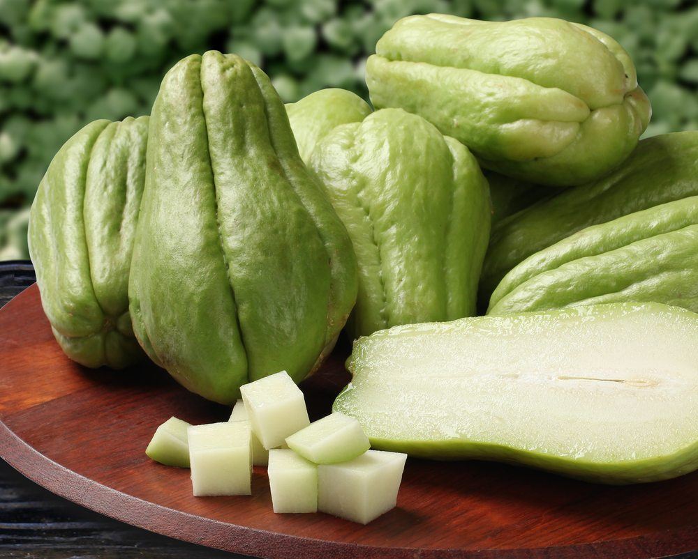 Fresh green chayote fruit. Most are whole, but one has been cut in half and there are wedges of the fruit as well. 