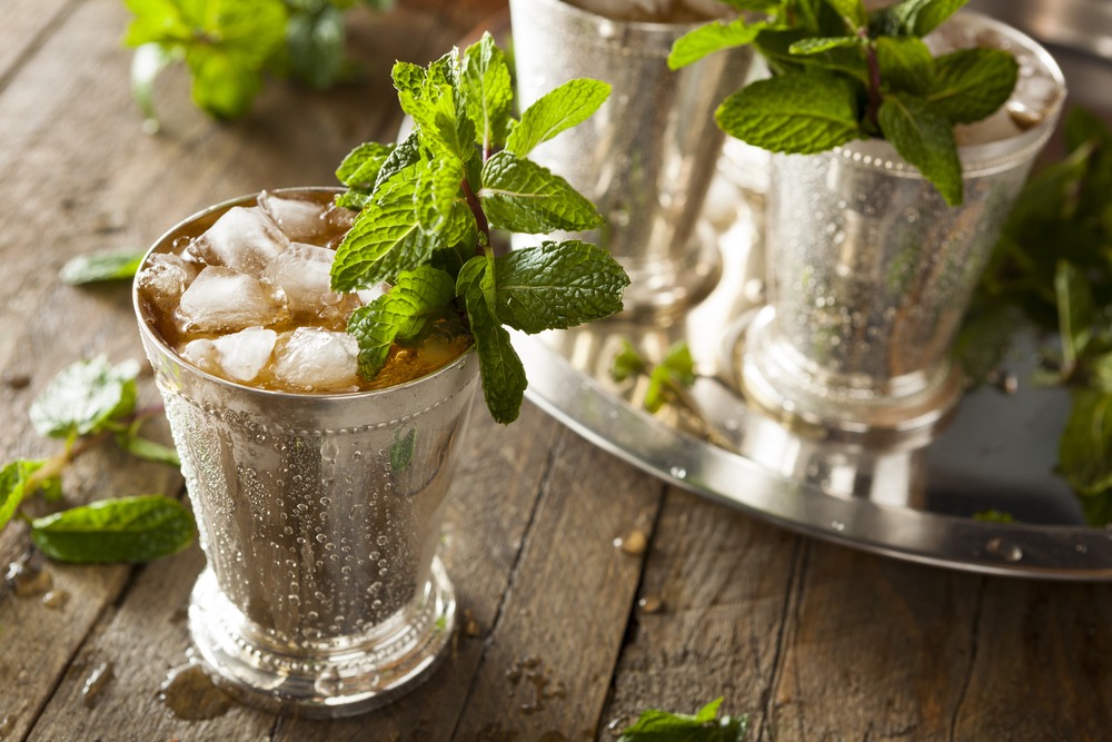 A mint julep in a stainless steel mug with ice and peppermint leaves, with a few more mint juleps on a tray