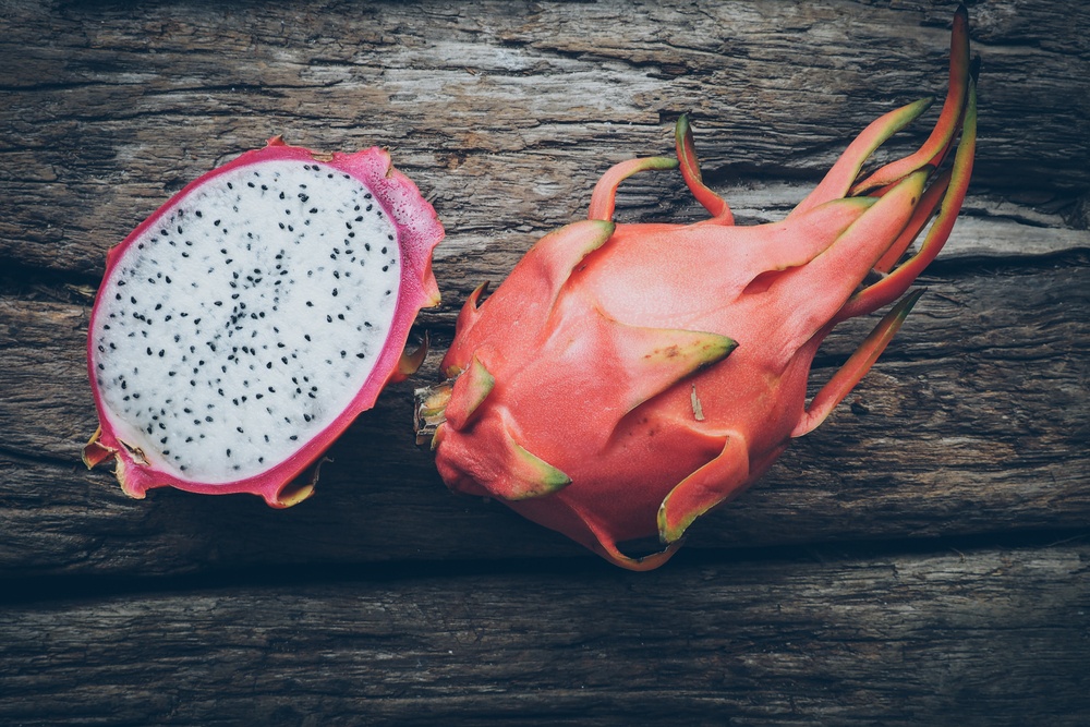 A whole dragon fruit on a wooden table, next to half a dragon fruit, where the white and black interior can be seen