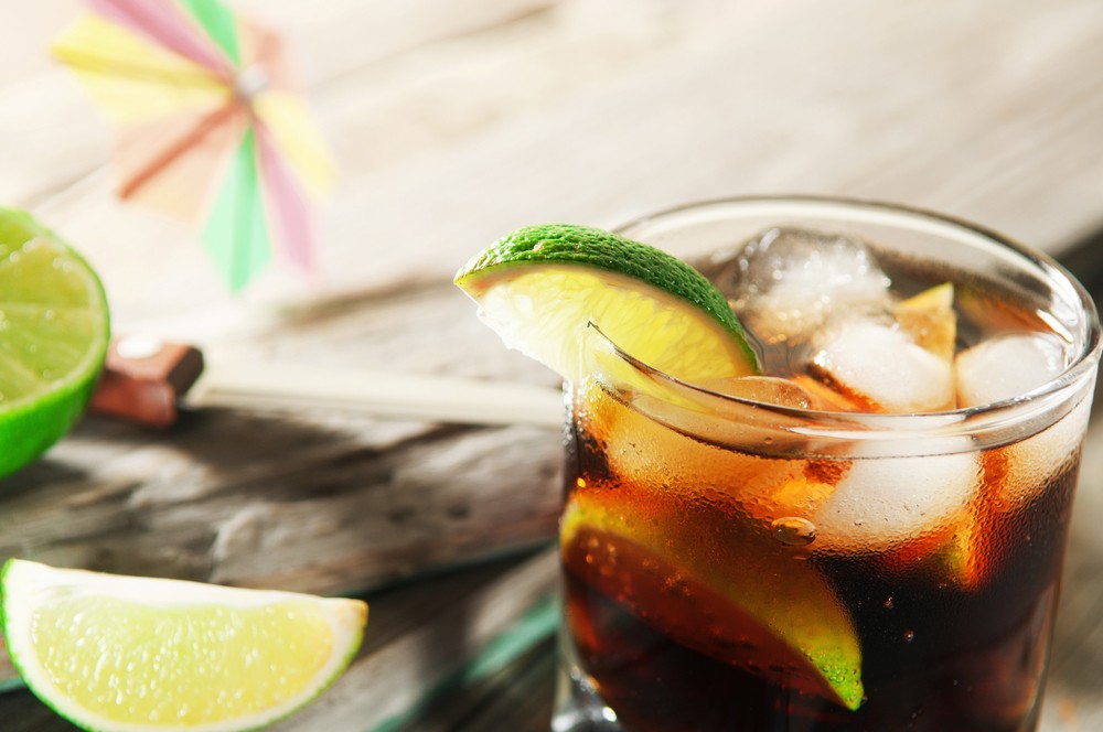 A glass of rum and coke with plenty of ice, which has been garnished with a lime wedge. There are also limes in the background. 