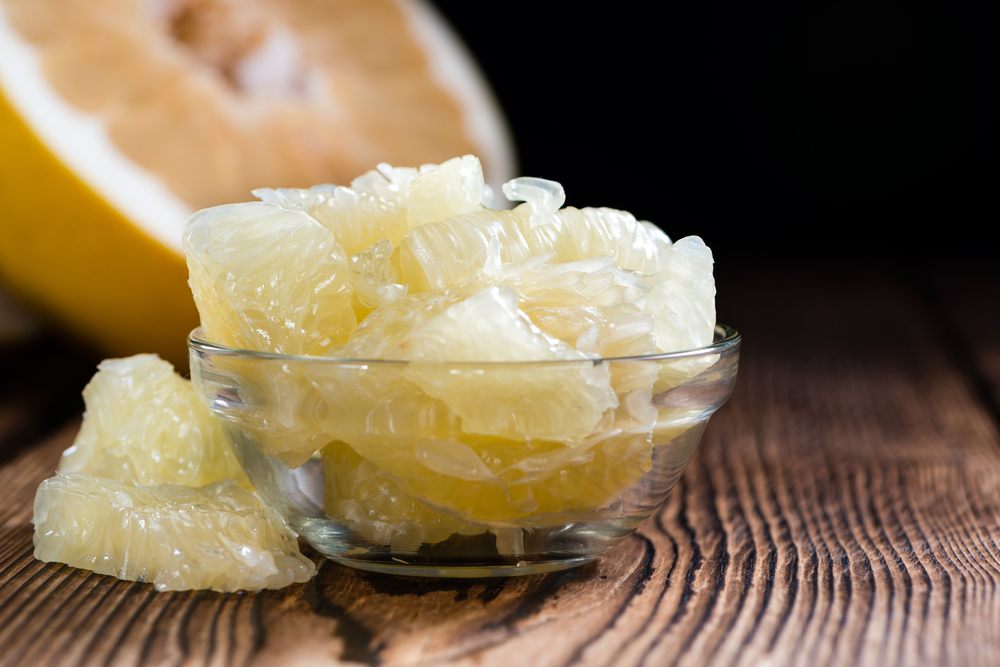 A glass bowl containing pomelo pieces with half a pomelo in the background