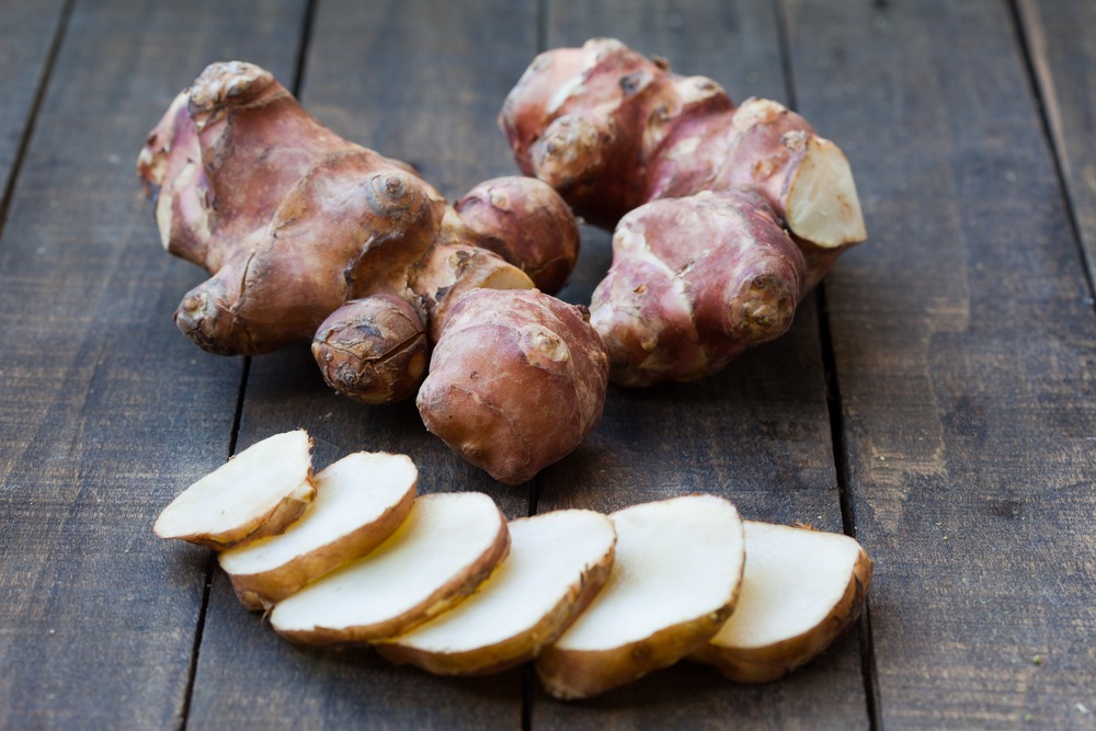 A couple of fresh sunchoke roots on a wooden table, with a sliced suncoke root in front of them