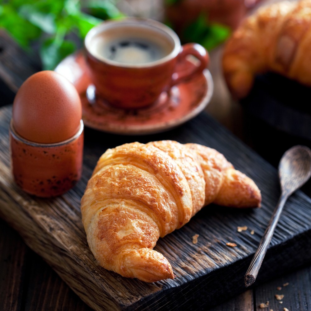 A wooden table with a croissant, a hard boiled egg, and a mug of coffee