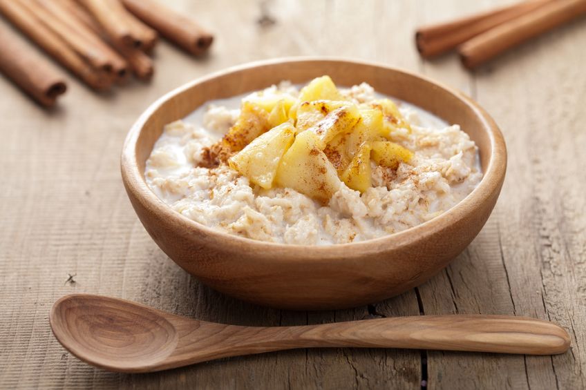 A wooden bowl filled with oatmeal, topped with cinnamon and apple, next to a wooden spoon