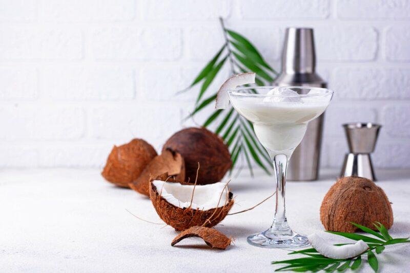 A coconut margarita cocktail on a white table next to some coconuts, a cocktail shaker, a jigger, some leaves, and some coconuts