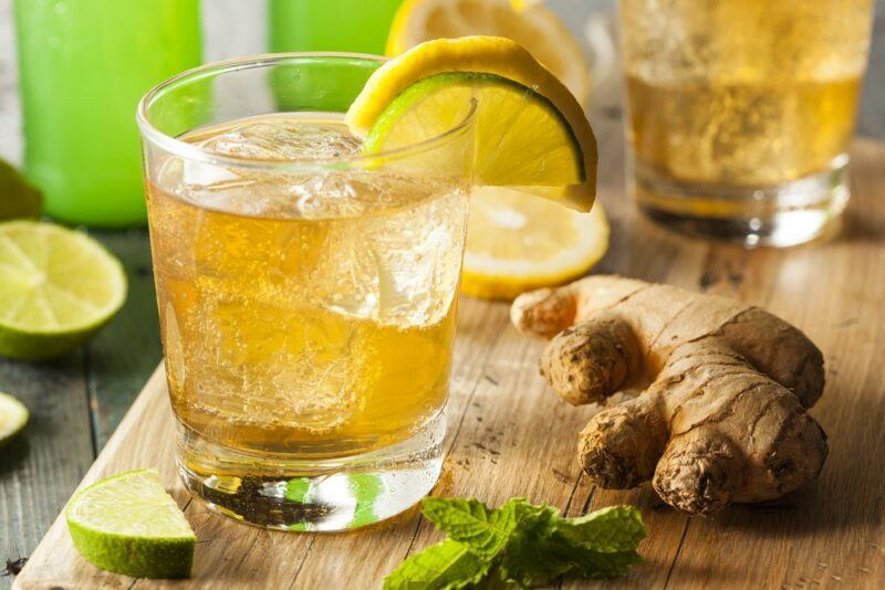 A Fireball, ginger beer, and apple cider cocktail on a board, next to a piece of ginger, some peppermint leaves, and lemon slices