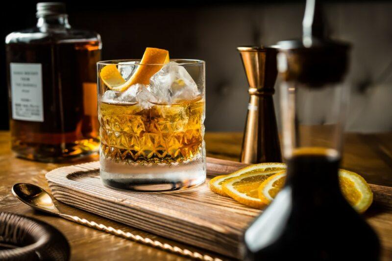 A wooden tray with an old fashioned or a Fireball old fashioned cocktail in a glass, next to a bottle of whiskey, some lemon slices, and bar tools