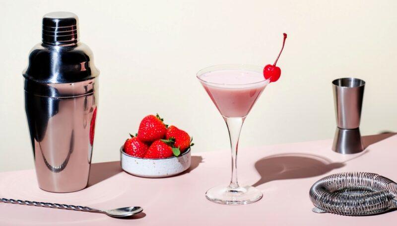 A light pink foxy lady cocktail, next to a cocktail jigger, a cocktail shaker, and other bar tools