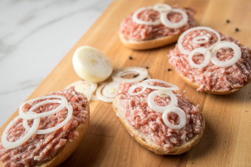 Four sliced bread rolls on a wooden board. Each has raw pork mince and sliced raw onion on top