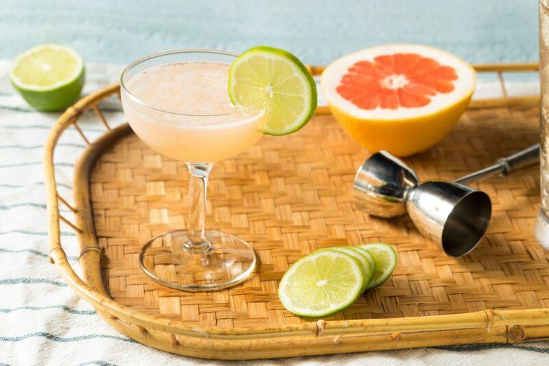 A wooden tray with a cocktail jigger, half a grapefruit, some lime slices, and a Hemingway daiquiri