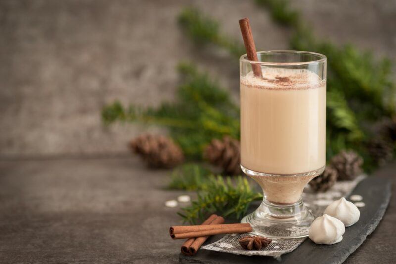 A single glass containing a Long Island coquito garnished with cinnamon, with winter decorations in the background and some cinnamon sticks