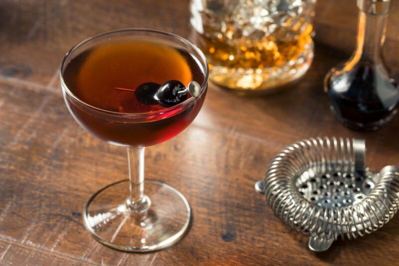 A Manhattan cocktail next to a cocktail strainer and a glass or decanter of bourbon