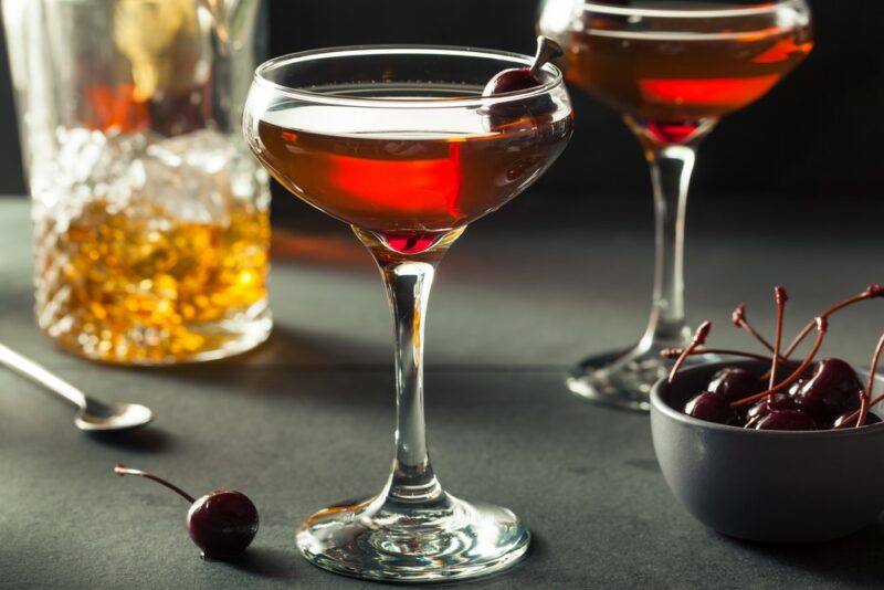 Two glasses containing a Manhattan fire cocktail, next to a glass of whiskey on a gray table