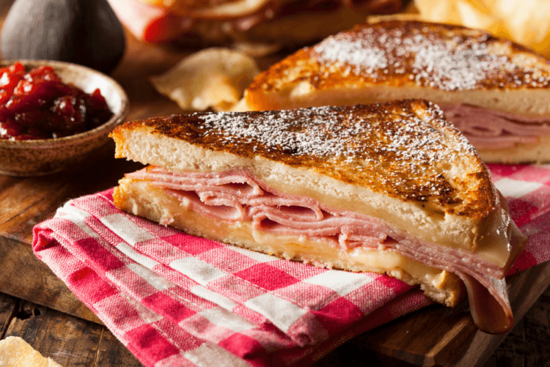 A wooden table with two halves of a Monte Cristo sandwich, a blue and white cloth, and a small bowl containing a condiment.