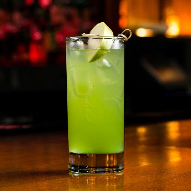 A single glass containing a Mountain Dew mojito cocktail with ice in front of an out of focus bar