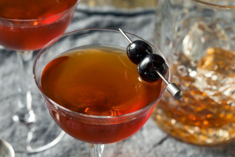 A Rob Roy cocktail in a coupe glass, garnished with two olives on a stick, with a second cocktail in the background