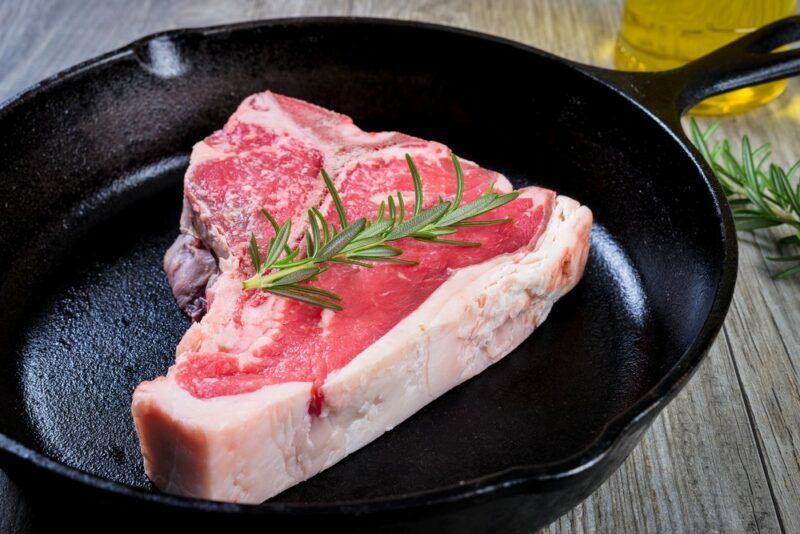 A cast iron fry pan containing a raw piece of T-bone steak and a small sprig of rosemary