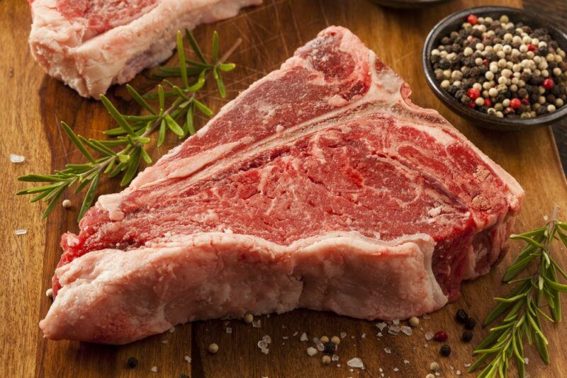 A wooden table with a piece of raw porterhouse steak (plus a second piece almost off camera), some fresh pepper, and some rosemary