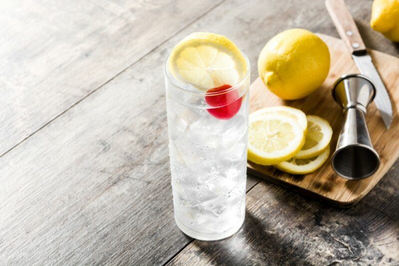 A wooden table with a Tom Collins cocktail in a tall glass, next to a whole lemon and some lemon slices