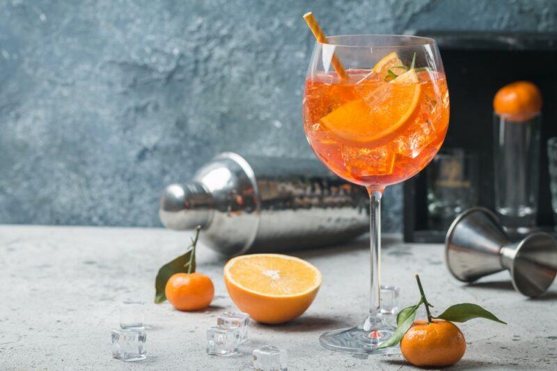 A large glass containing an Aperol spritz cocktail with a straw and orange wedges, on a table with half an orange and small mandarins, with a cocktail shaker in the background