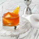 A single glass on a light wooden board, containing an orange Wilde Oscar old fashioned cocktail