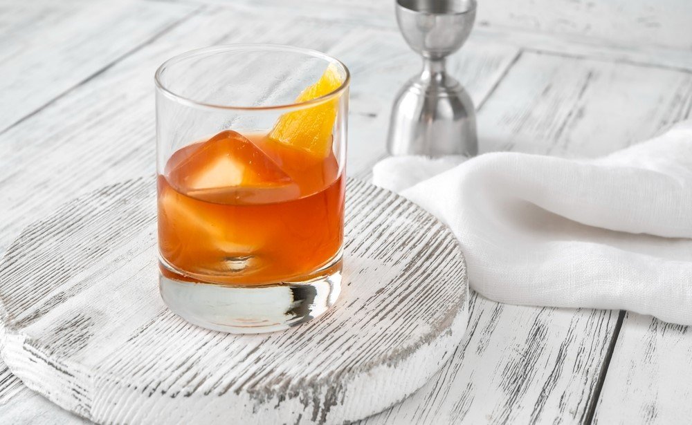 A single glass on a light wooden board, containing an orange Wilde Oscar old fashioned cocktail