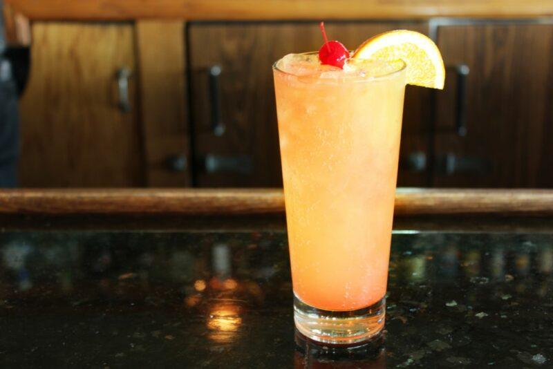 A tall glass containing a Bahama mama cocktail with a glace cherry and lemon wedge, on a counter