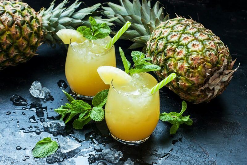 Two glasses containing a pineapple baja tiki cocktail with pineapples in the background