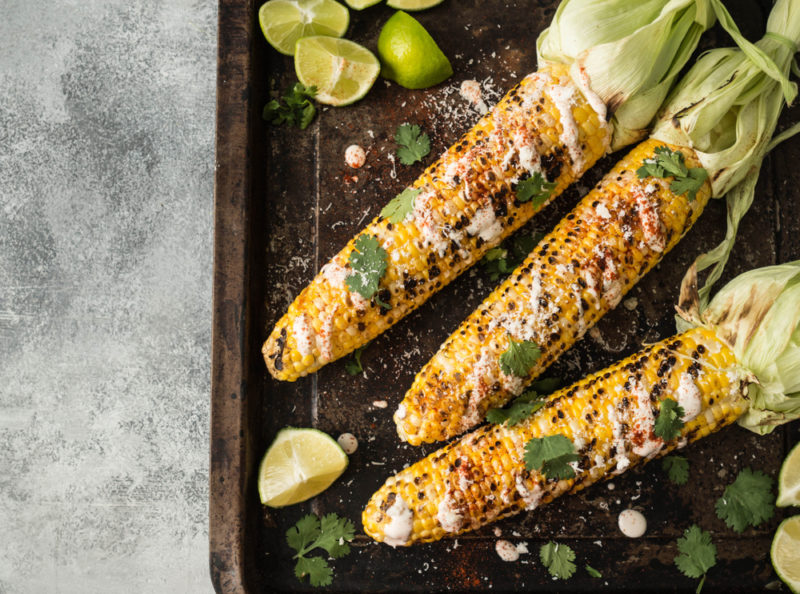 A baking tray with three cobs of corn that have been grilled and sprinkled with sauce. There are some limes and a few herbs on the tray too