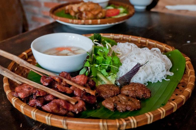 A bamboo dish with a street food called bun cha, along with noodles, broth, and chopsticks, with a second similar tray in the background