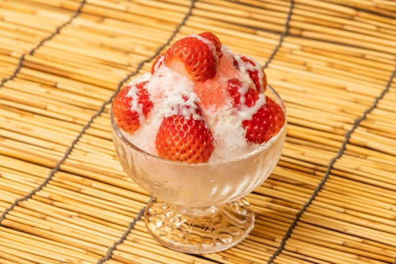 A glass container on a bamboo mat, with a banana berry snow cone, condensed milk, and fresh strawberries