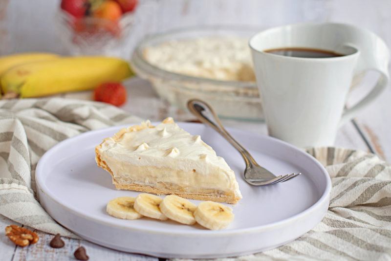 A white dish containing a banana cream pie and sliced bananas, next to a mug of coffee and ingredients for the pie