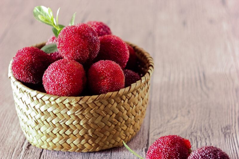 A basket containing fresh sweet and sour yangmei fruits on a wooden table, with a few fruits on the table itself