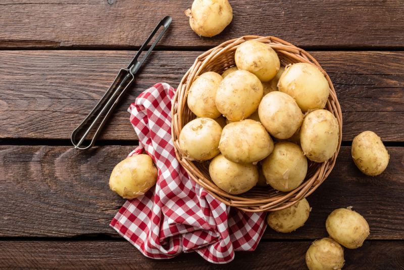 A basket on a red and wite cloth, filled with raw potatoes, next to a potato peeler