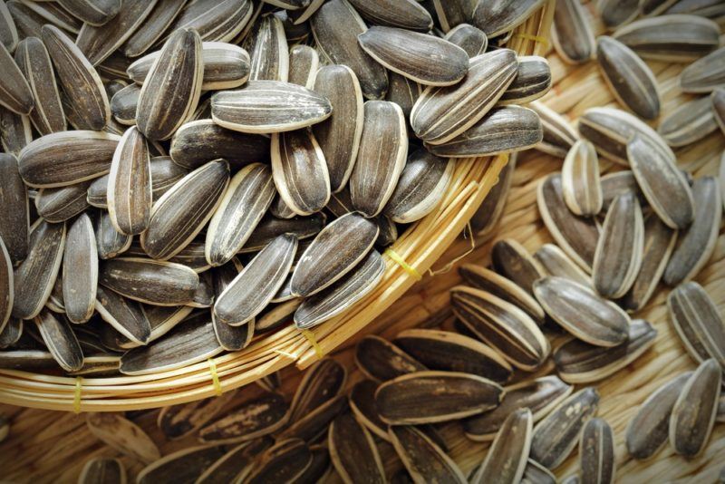 A brown basket that contains pumpkin seeds, with more seeds on the table