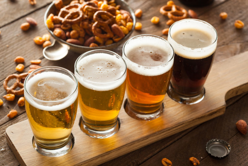 A wooden tray with four glasses of beer of different types, along with bar snacks scattered in the background