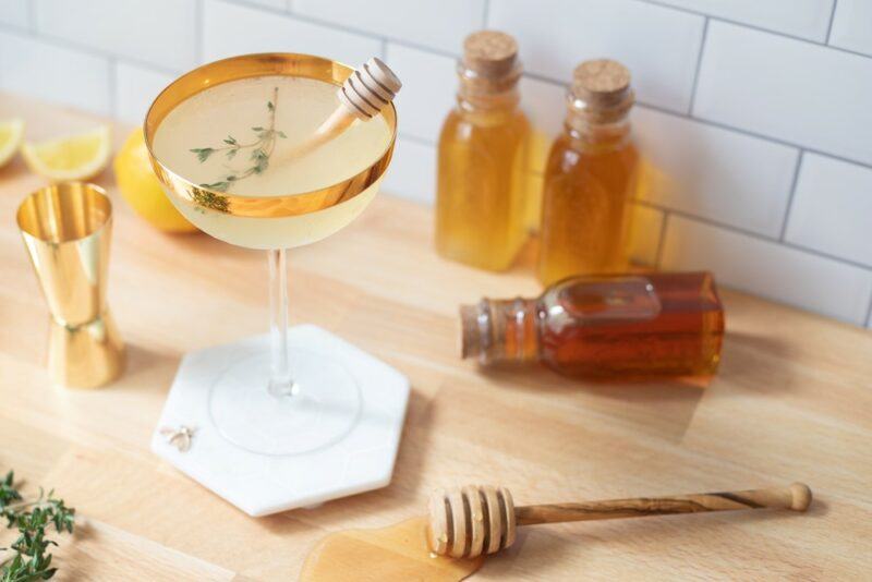 A table with a bee's tease cocktail next to small bottles of honey and a honey stirrer