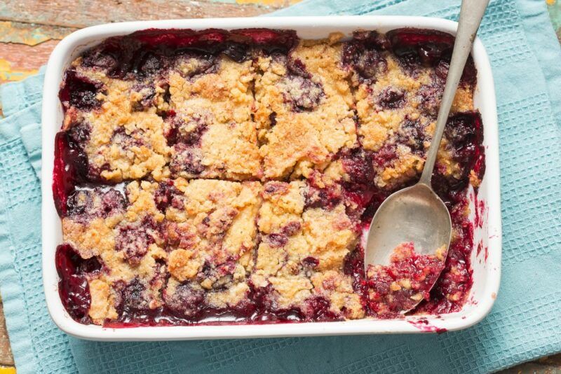 A large white dish containing a berry cream cheese dump cake that has been cut into spices, with a spoon