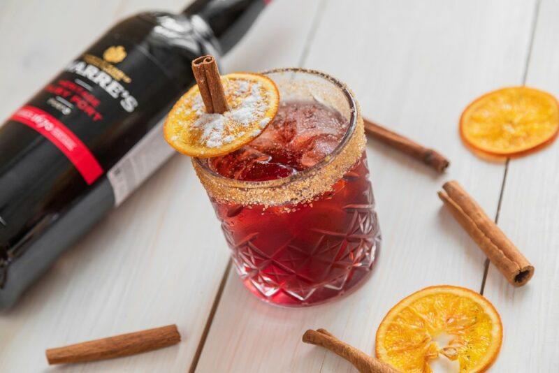 A light wooden table containing a bishop cocktail next to cinnamon sticks, fruit slices, and a bottle of port