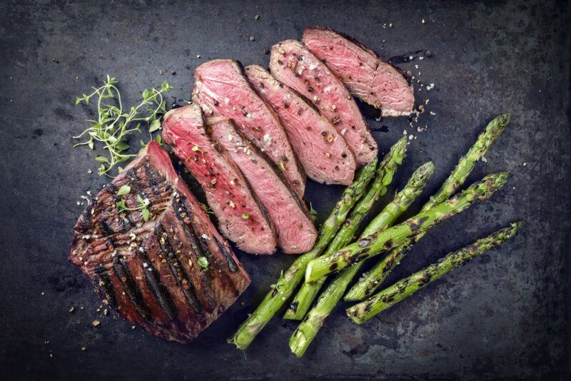 A black background with a large piece of sirloin tip steak that has been sliced into thin pieces, next to asparagus spears and some sprigs of rosemary