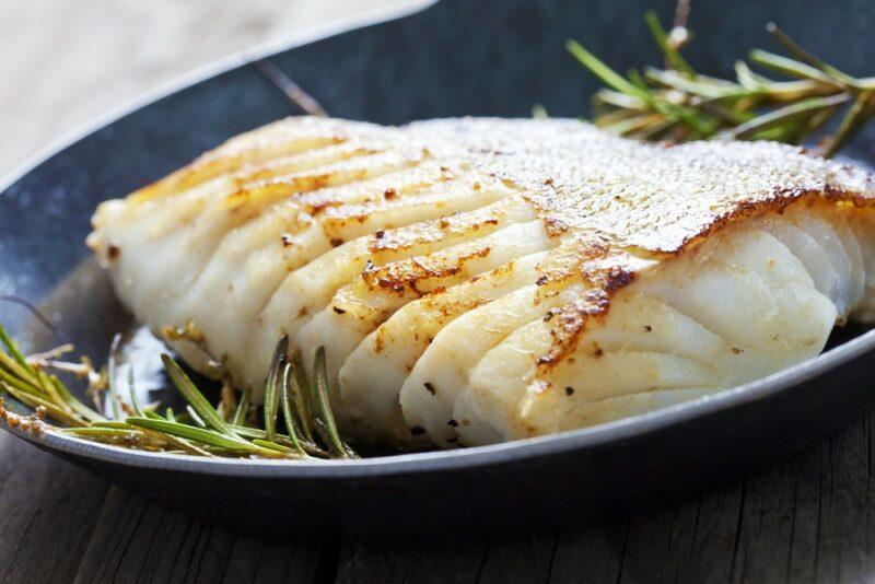 A dark colored bowl containing cooked white fish and some sprigs of rosemary