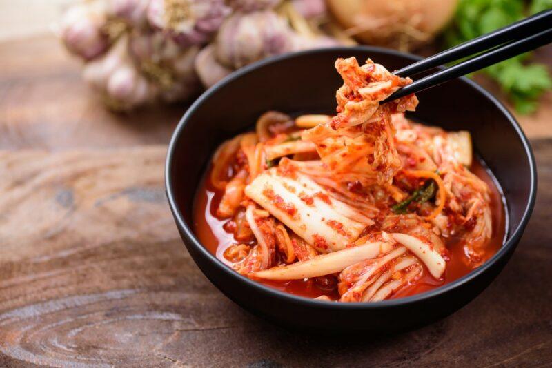 A black bowl on a wooden table, containing kimchi and plenty of juice. A small amount is being picked up using chopsticks.