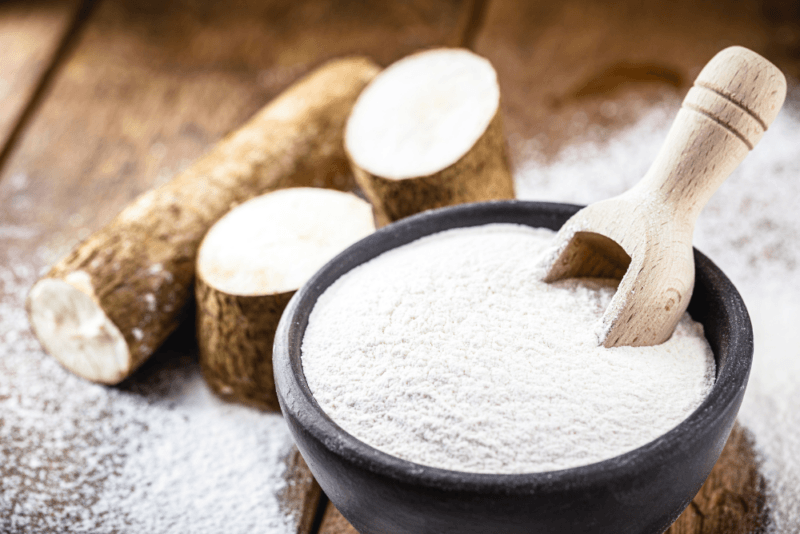 A wooden table with a few cassava roots and a large black bowl containing cassava flour with a wooden scoop