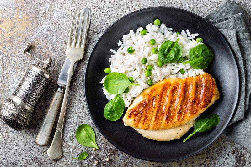 A black dish of rice with greens and chicken, next to a knife, fork and pepper grinder