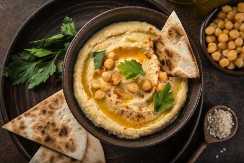 A brown bowl filled with hummus, with pita bread sticking out of it and some more pita bread and some chickpeas next to it